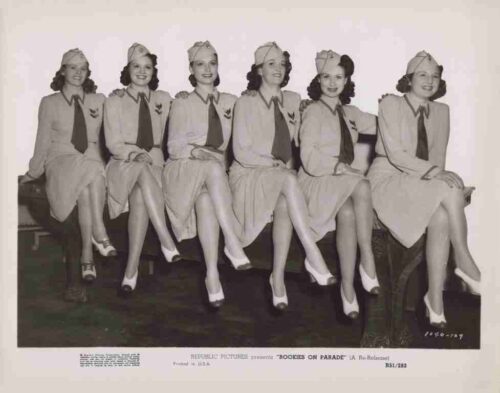 Seven women in matching 1940s-style uniforms sit in a row on a bench, smiling and crossing one leg over the other.