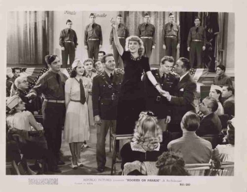 A woman in a dark dress raises her arm on a stage as soldiers and an audience look on in a black-and-white film scene.