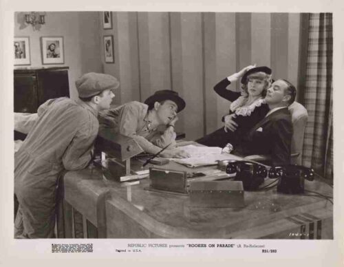 Vintage film publicity photo: a suited man sits at a desk with a woman on his lap as another woman holds a child, while two men lean over the desk to look at papers on the desk.