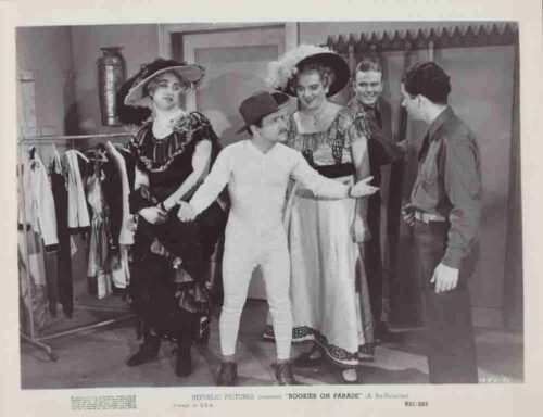 Group of performers in vintage costumes backstage, with a central mime-like boy in white extending his arms as others smile nearby.
