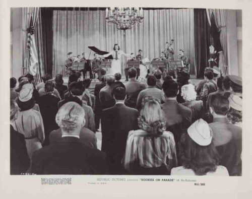 Black-and-white photo of a mid-20th-century concert: a singer with arms outstretched performs on stage with an accompanying band, while a standing audience watches.