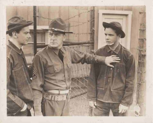 Three men in vintage military attire stand by a barbed-wire fence; the central figure rests a hand on the shoulder of the man to his right.