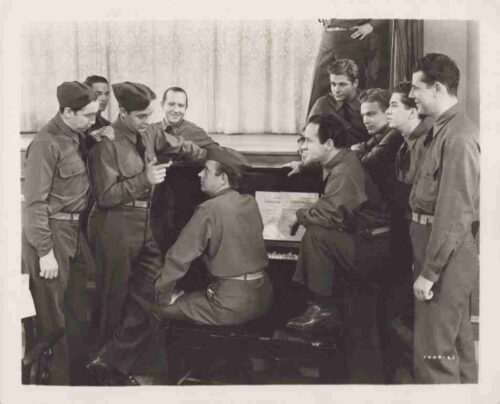 Group of soldiers in uniform gathered around a piano, with one seated at the keyboard as others lean in and smile.